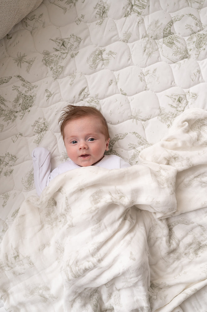 Close-up of a baby beneath a matching safari-print blanket on the quilted playmat, with pale green animal illustrations.