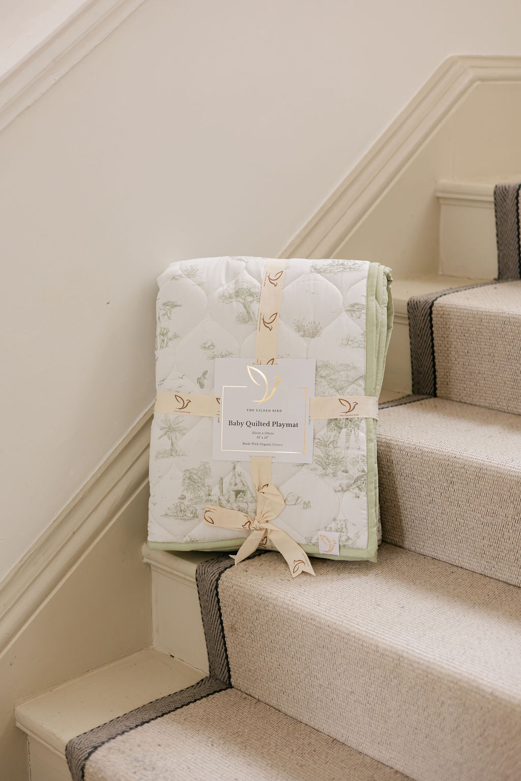 Safari Toile quilted playmat folded and tied with branded ribbon on a carpeted stair, showing its label and green edging.