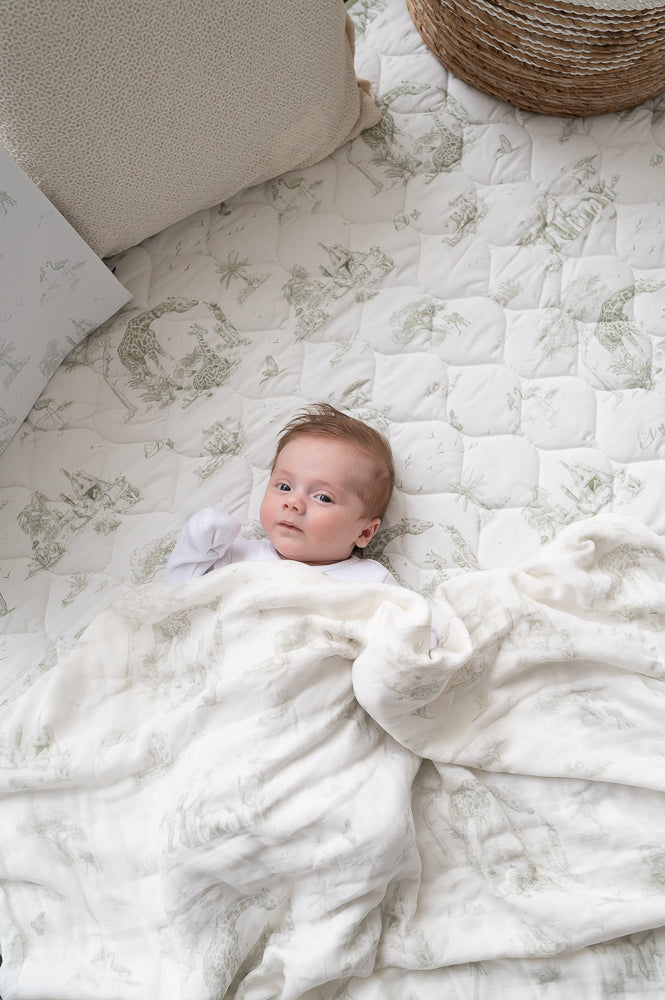 Baby lying on the Safari Toile quilted playmat beneath a matching animal-print blanket, beside a cushion and woven basket.