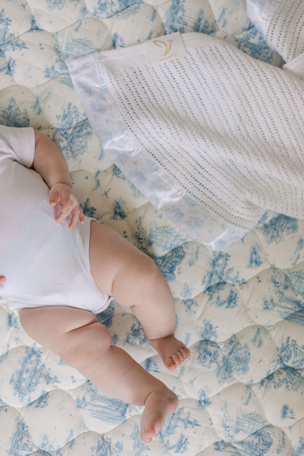 Close-up of a baby’s legs on the blue toile quilted playmat beside a white knitted cellular baby blanket.