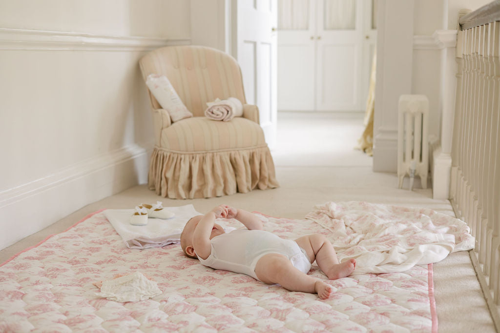Baby lying on the Spring Toile Pink quilted playmat beside a crib, with a matching blanket on the mat.
