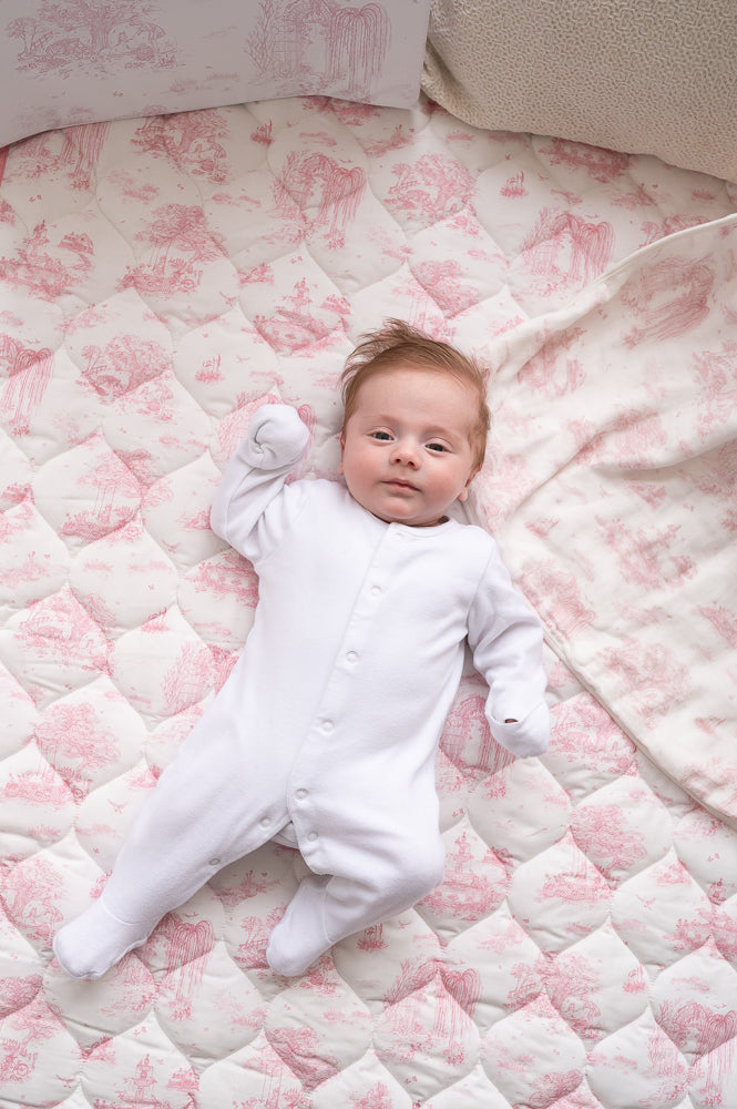 Baby in a white sleepsuit lying on the pink toile quilted playmat beside a matching patterned blanket.